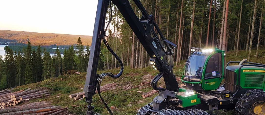 Grønn skogsmaskin på en åsside med tømmerstokker og en innsjø i bakgrunnen