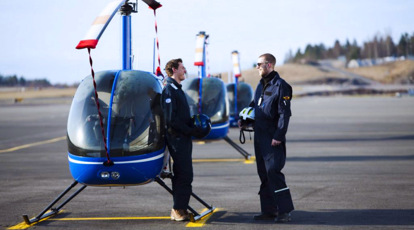 To personer i flydresser står ved blå helikoptre på en rullebane og holder hjelmer