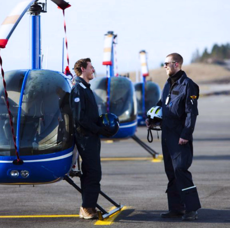 To personer i flydresser står ved blå helikoptre på en rullebane og holder hjelmer