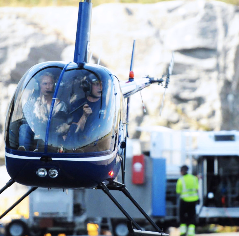 Blått og hvitt helikopter i luften med to personer i cockpit og fjell i bakgrunnen