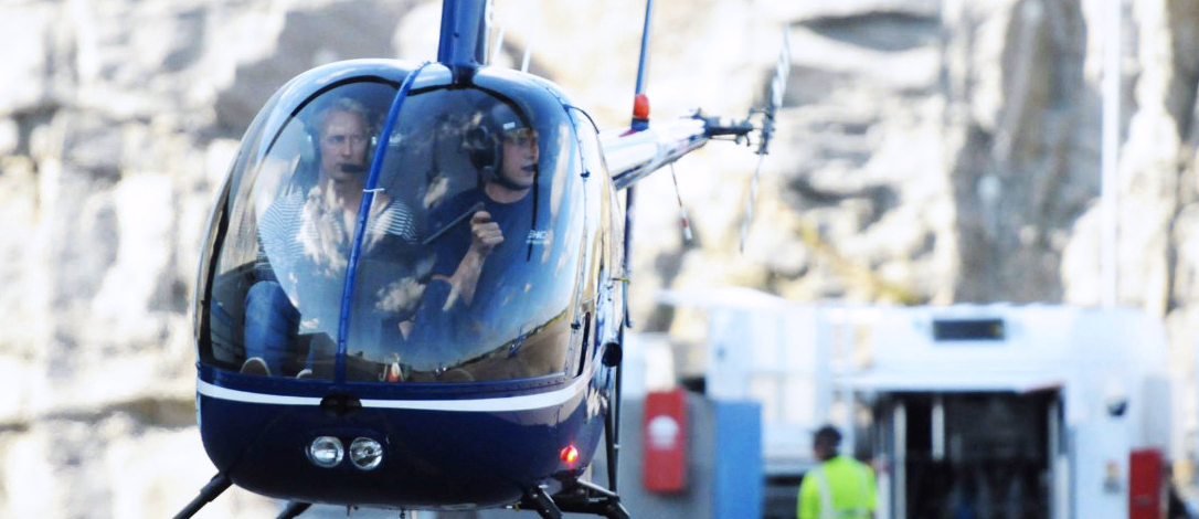 Blått og hvitt helikopter i luften med to personer i cockpit og fjell i bakgrunnen