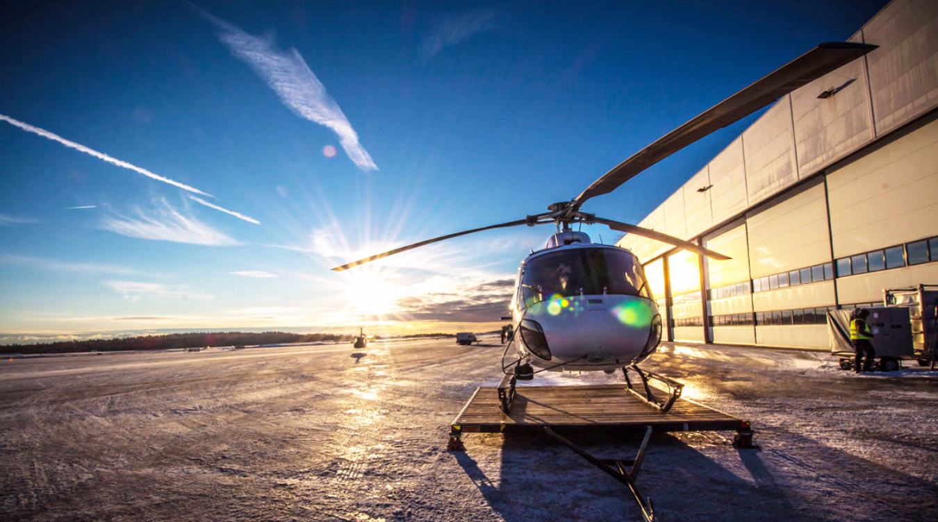 Sølvfarget helikopter på snødekket bakke ved en hvit bygning i lav sol