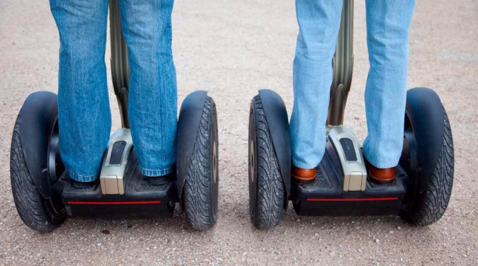 To personer i blå jeans på hver sin ståhjuling med store svarte hjul på lys grus