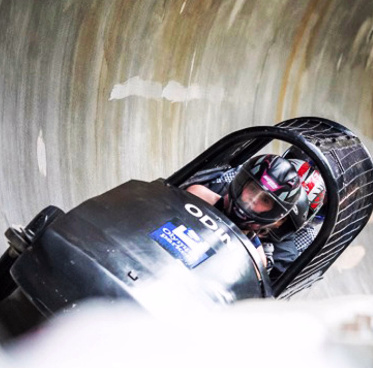 Svart bobslede med person i hjelm i fart gjennom en svingete iskanal