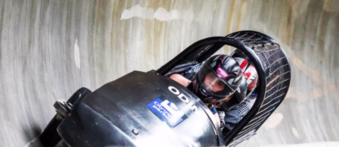Svart bobslede med person i hjelm i fart gjennom en svingete iskanal