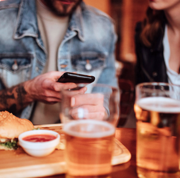 To personer ved et bord med burgere på trebrett, to glass øl og en mobiltelefon