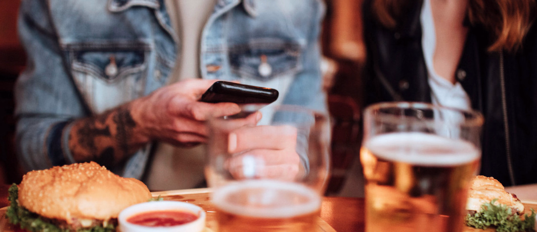 To personer ved et bord med burgere på trebrett, to glass øl og en mobiltelefon