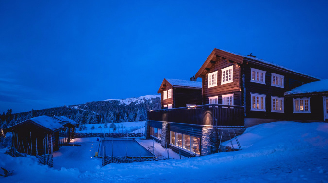 Stor trehytte med lys i vinduene i et snødekt kveldslandskap
