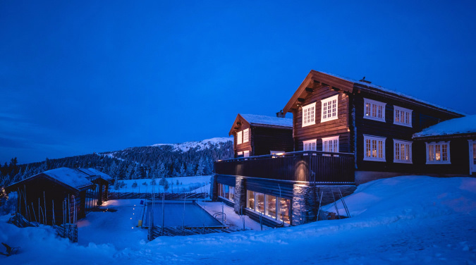 Stor trehytte med lys i vinduene i et snødekt kveldslandskap