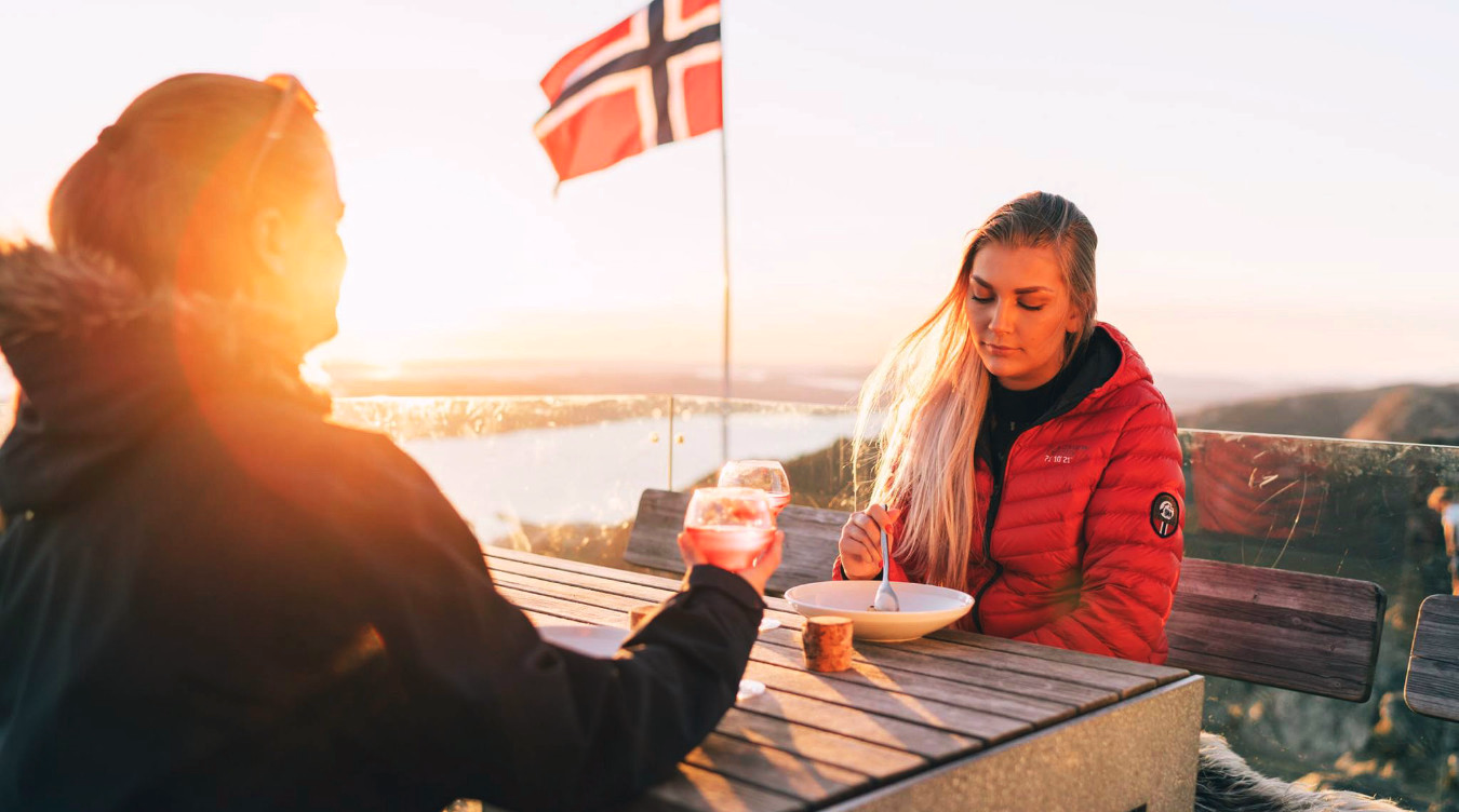 To personer sitter utendørs ved et bord med mat og drikke og et norsk flagg i bakgrunnen