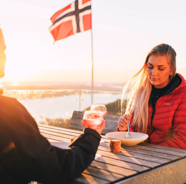 To personer sitter utendørs ved et bord med mat og drikke og et norsk flagg i bakgrunnen
