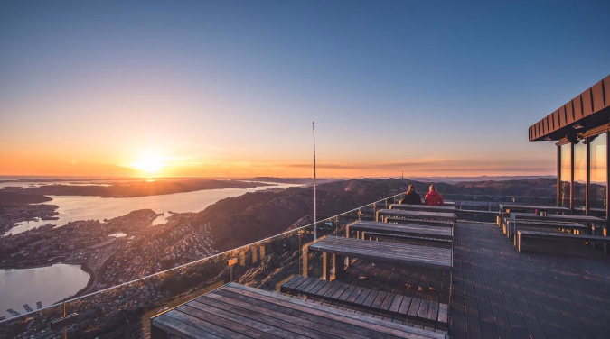 To personer på en trebenk ser ut over et landskap med vann og åser i solnedgang