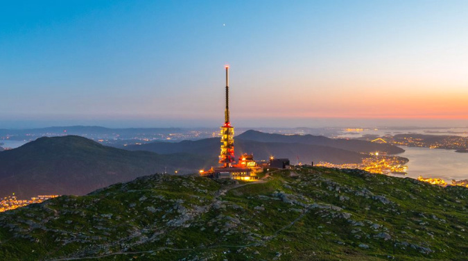 Høyt tårn med røde lys på en grønn ås over en opplyst by ved vannet