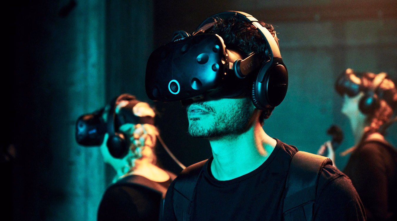 Person med svart VR-headset og hodetelefoner i blågrønt lys med andre i bakgrunnen