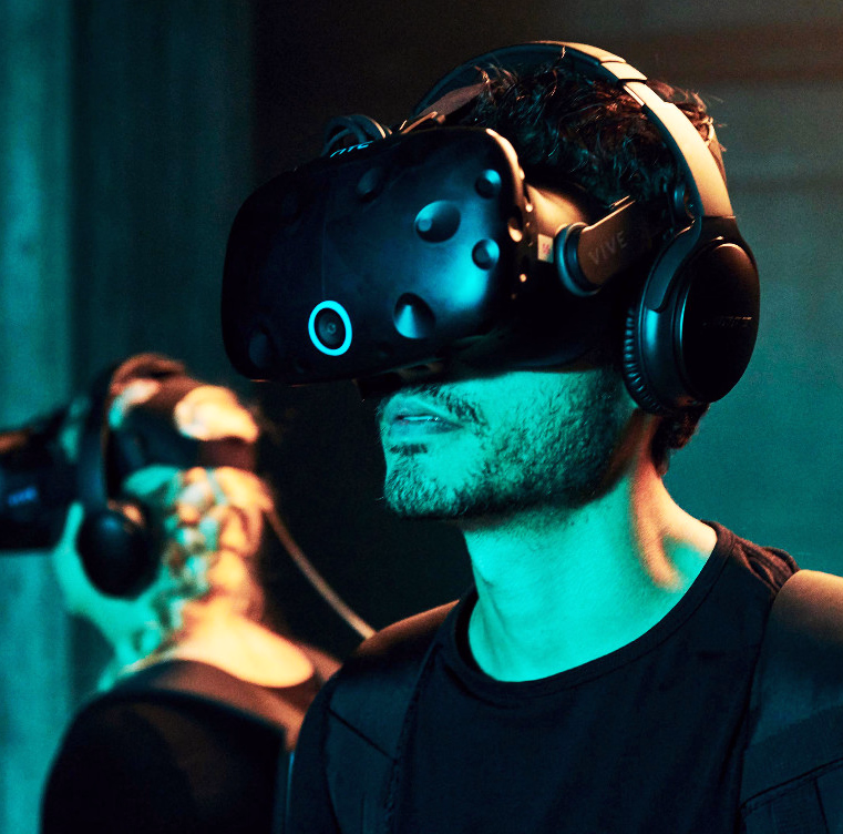 Person med svart VR-headset og hodetelefoner i blågrønt lys med andre i bakgrunnen