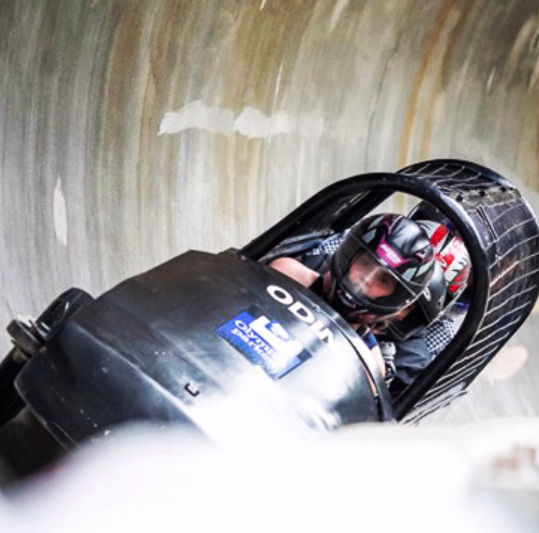 To personer i en svart bobslede med teksten ODIN på en isbane