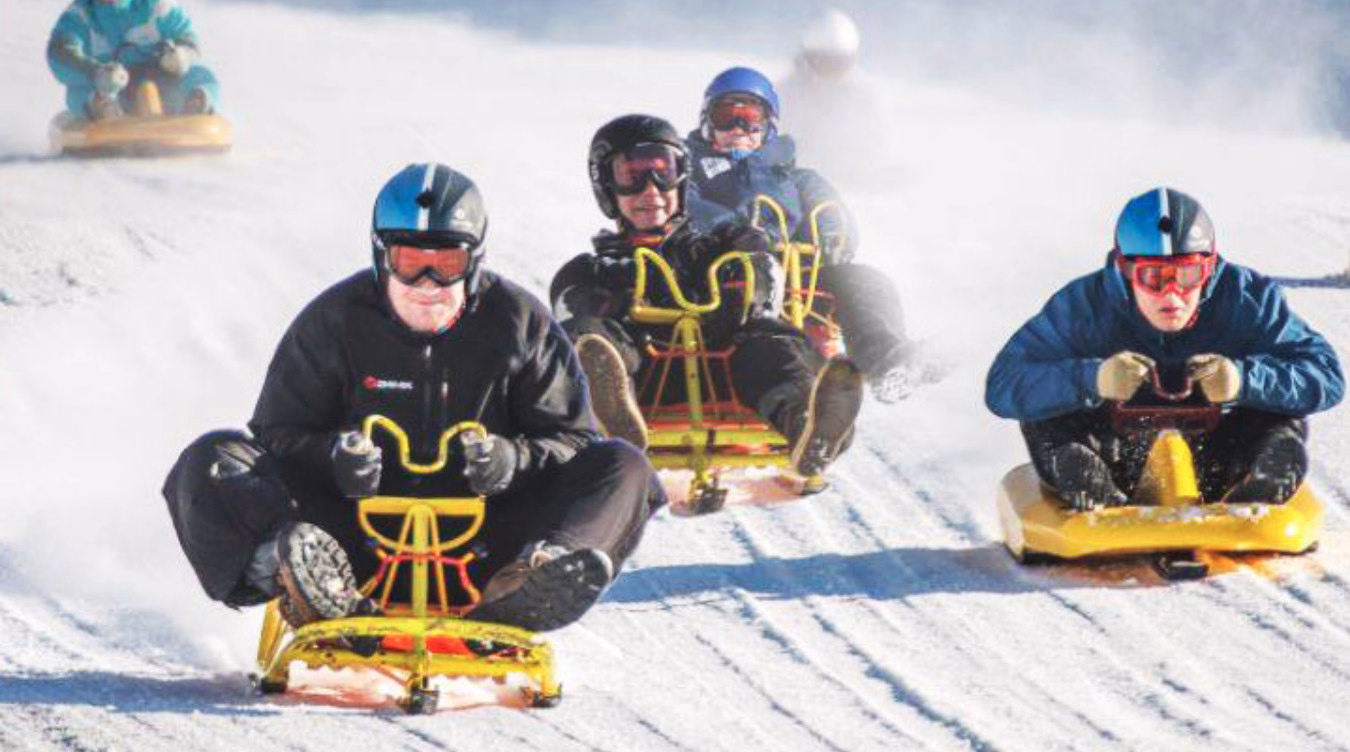 Personer med hjelm aker ned en snøbakke på gule kjelker med snøsprut