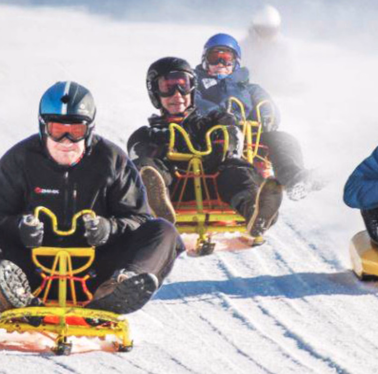 Personer med hjelm aker ned en snøbakke på gule kjelker med snøsprut