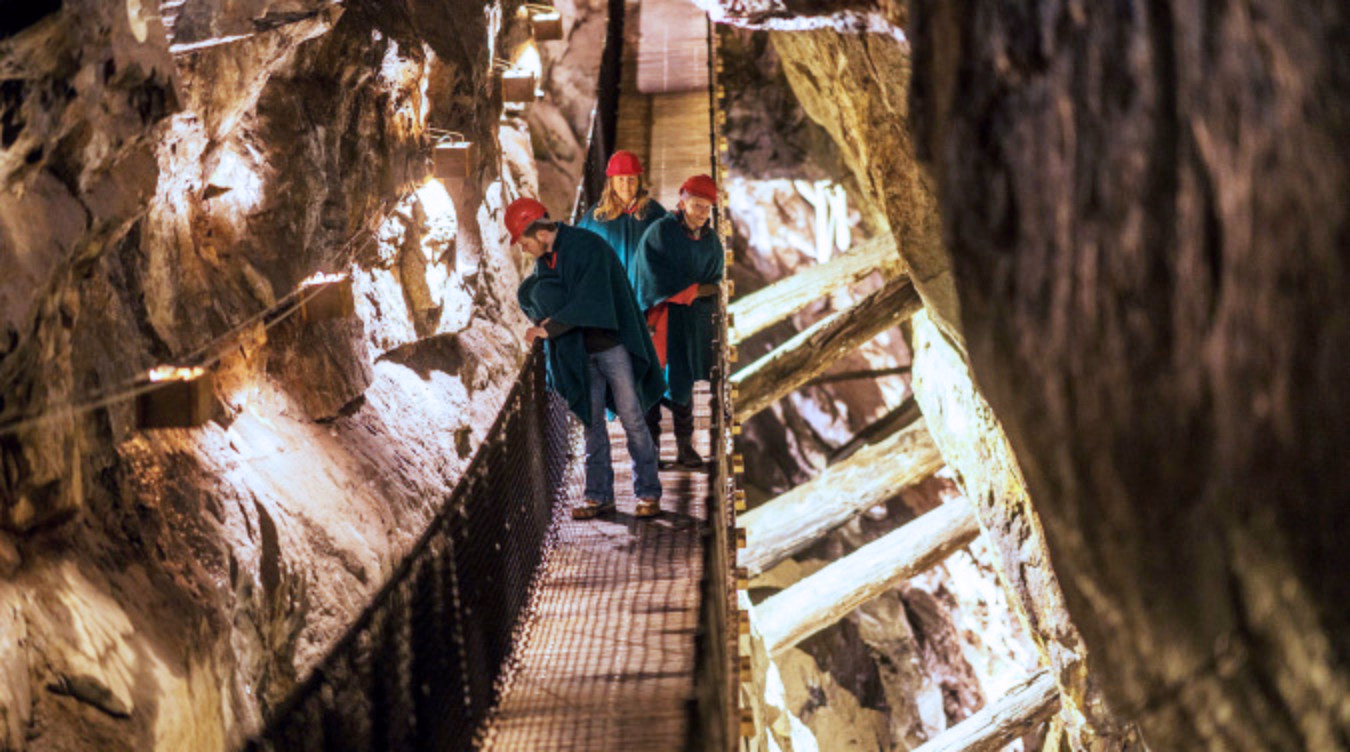 Tre personer med røde hjelmer og blå kapper på en trebro i en opplyst grotte