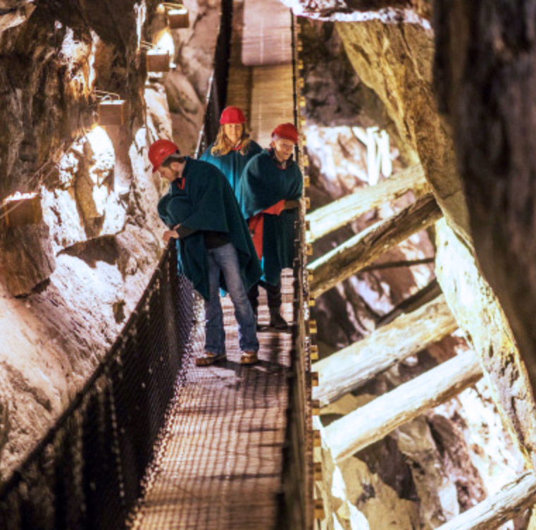 Tre personer med røde hjelmer og blå kapper på en trebro i en opplyst grotte