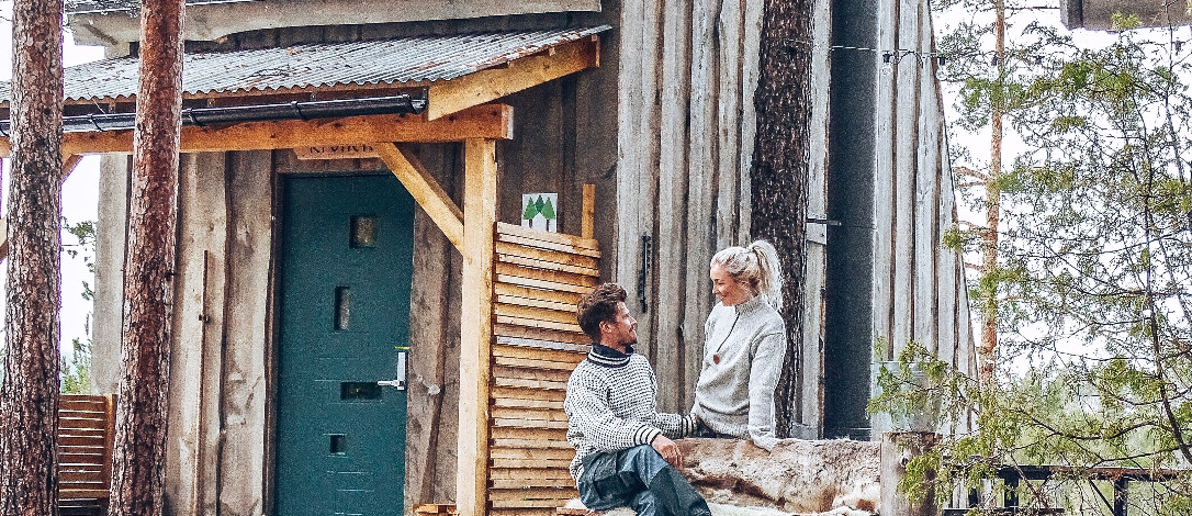 To personer sitter på en benk med skinnfell utenfor en trehytte med grønn dør