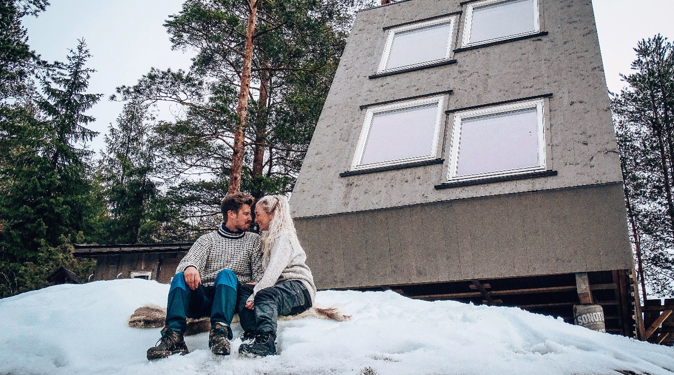 Par i strikkegensere sitter i snøen foran en grå A-ramme hytte