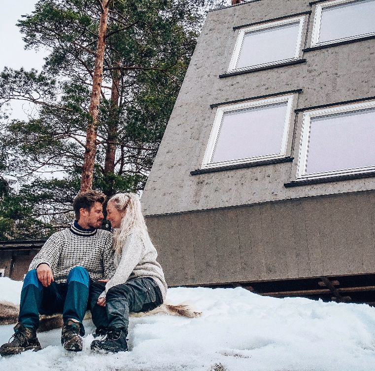 Par i strikkegensere sitter i snøen foran en grå A-ramme hytte
