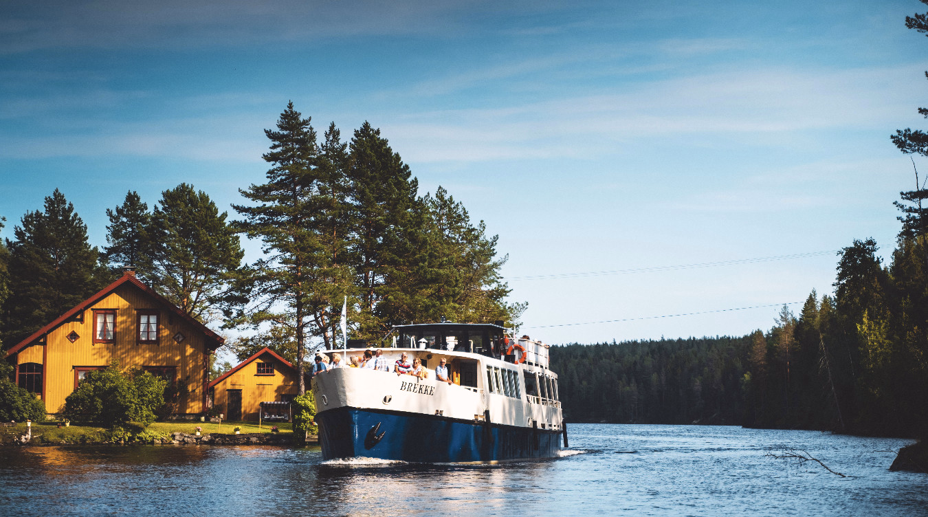 Hvit og blå båt med navnet BREKKE og folk om bord på en elv ved et gult hus og skog