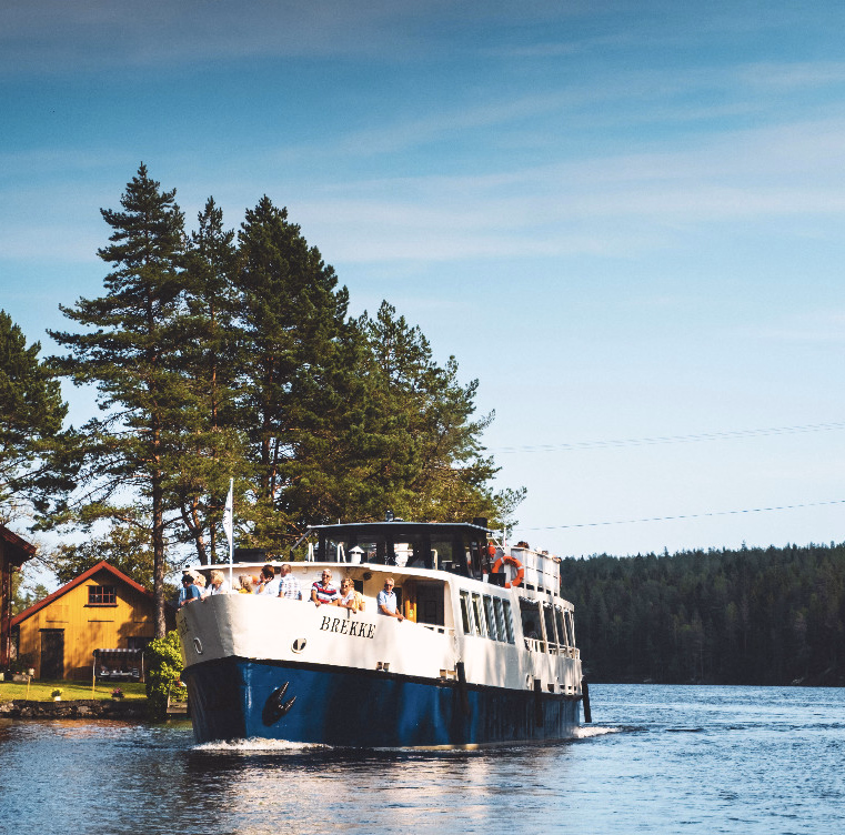 Hvit og blå båt med navnet BREKKE og folk om bord på en elv ved et gult hus og skog