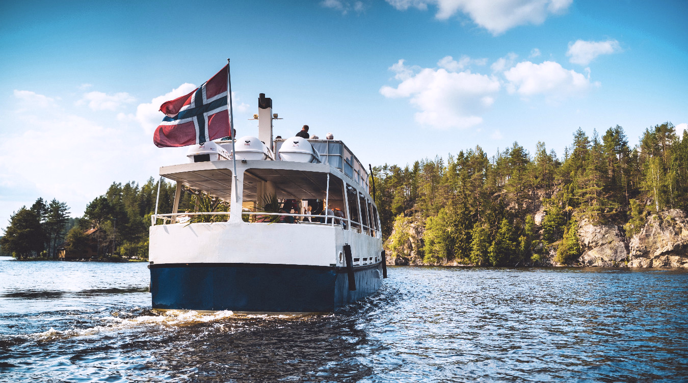 Hvit og blå båt med passasjerer på en innsjø foran skogkledde klipper