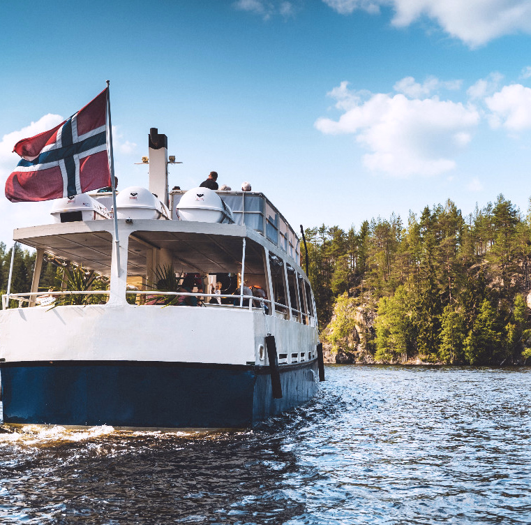 Hvit og blå båt med passasjerer på en innsjø foran skogkledde klipper
