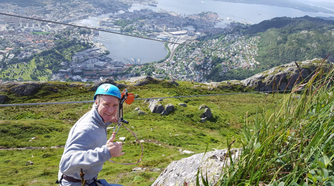 Person med blå hjelm på zipline gir tommel opp med utsikt over by og landskap