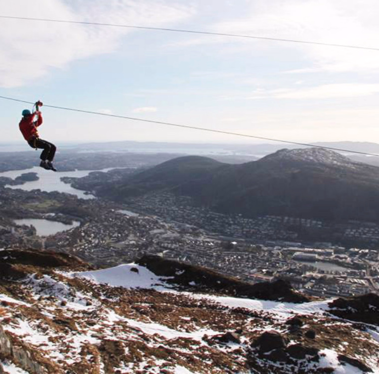Person i rød jakke og blå hjelm kjører zipline over fjellandskap med utsikt mot en by