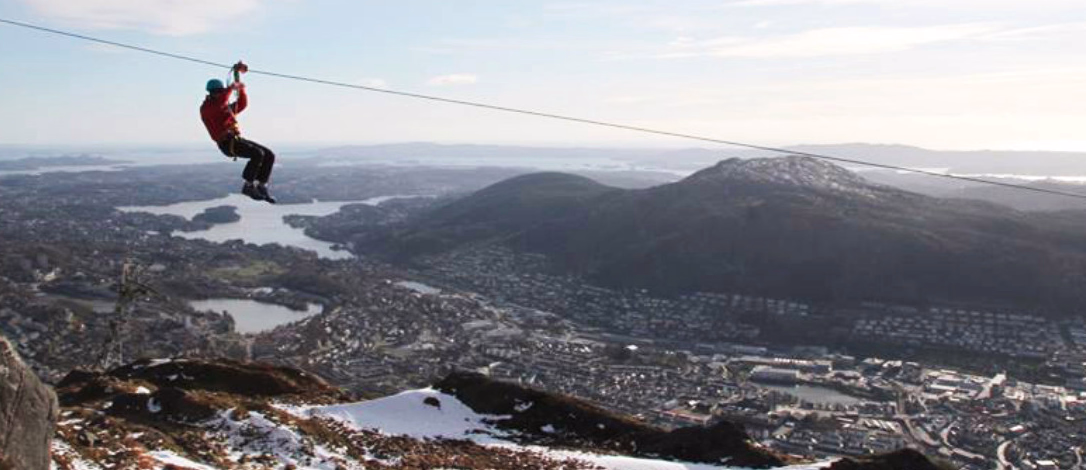 Person i rød jakke og blå hjelm kjører zipline over fjellandskap med utsikt mot en by