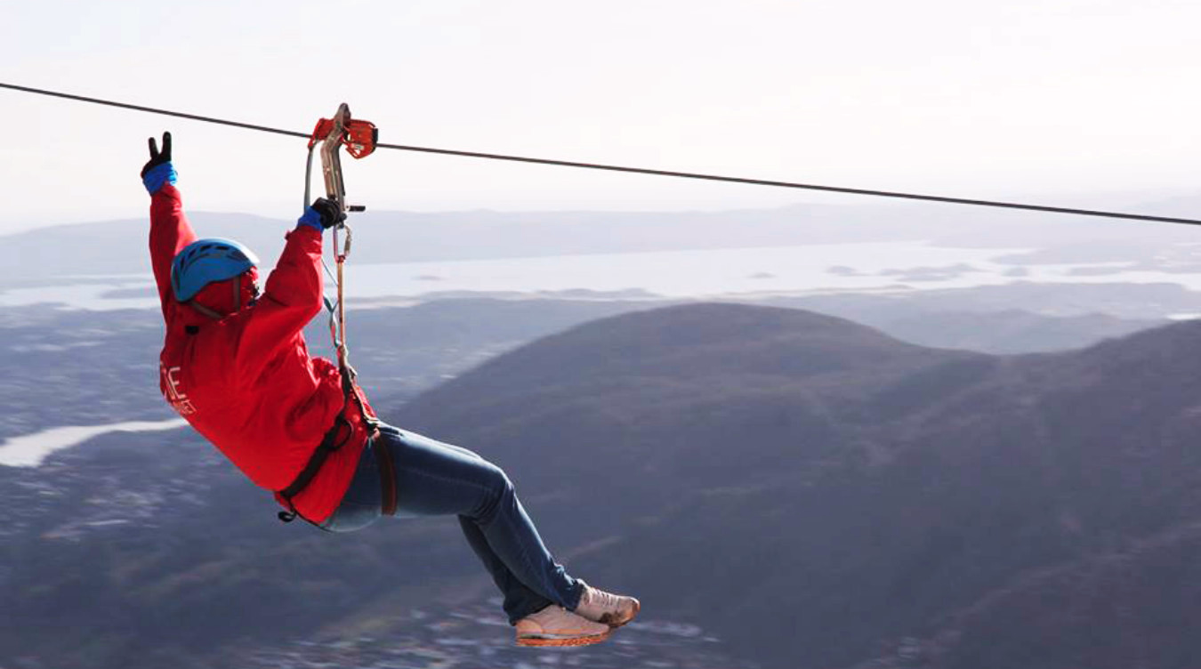 Person i rød jakke og blå hjelm som kjører zipline og viser fredstegn