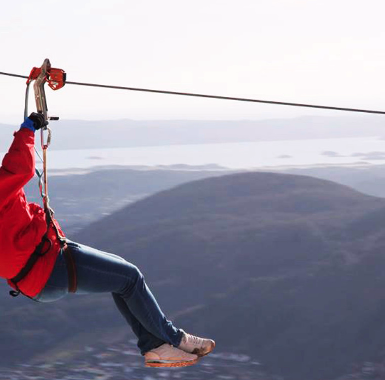 Person i rød jakke og blå hjelm som kjører zipline og viser fredstegn