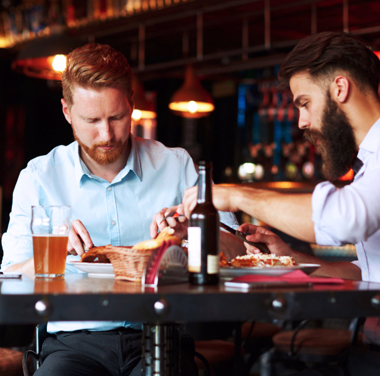To menn spiser pizza og drikker øl i en restaurant med hengende lamper