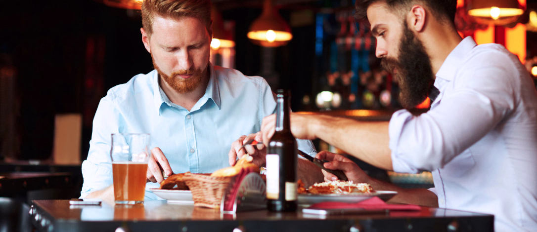 To menn spiser pizza og drikker øl i en restaurant med hengende lamper