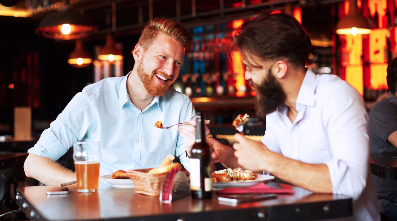 To menn som smiler og spiser ved et restaurantbord med mat og drikke