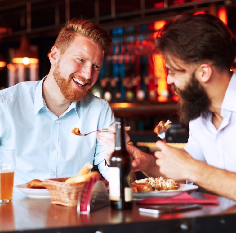 To menn som smiler og spiser ved et restaurantbord med mat og drikke