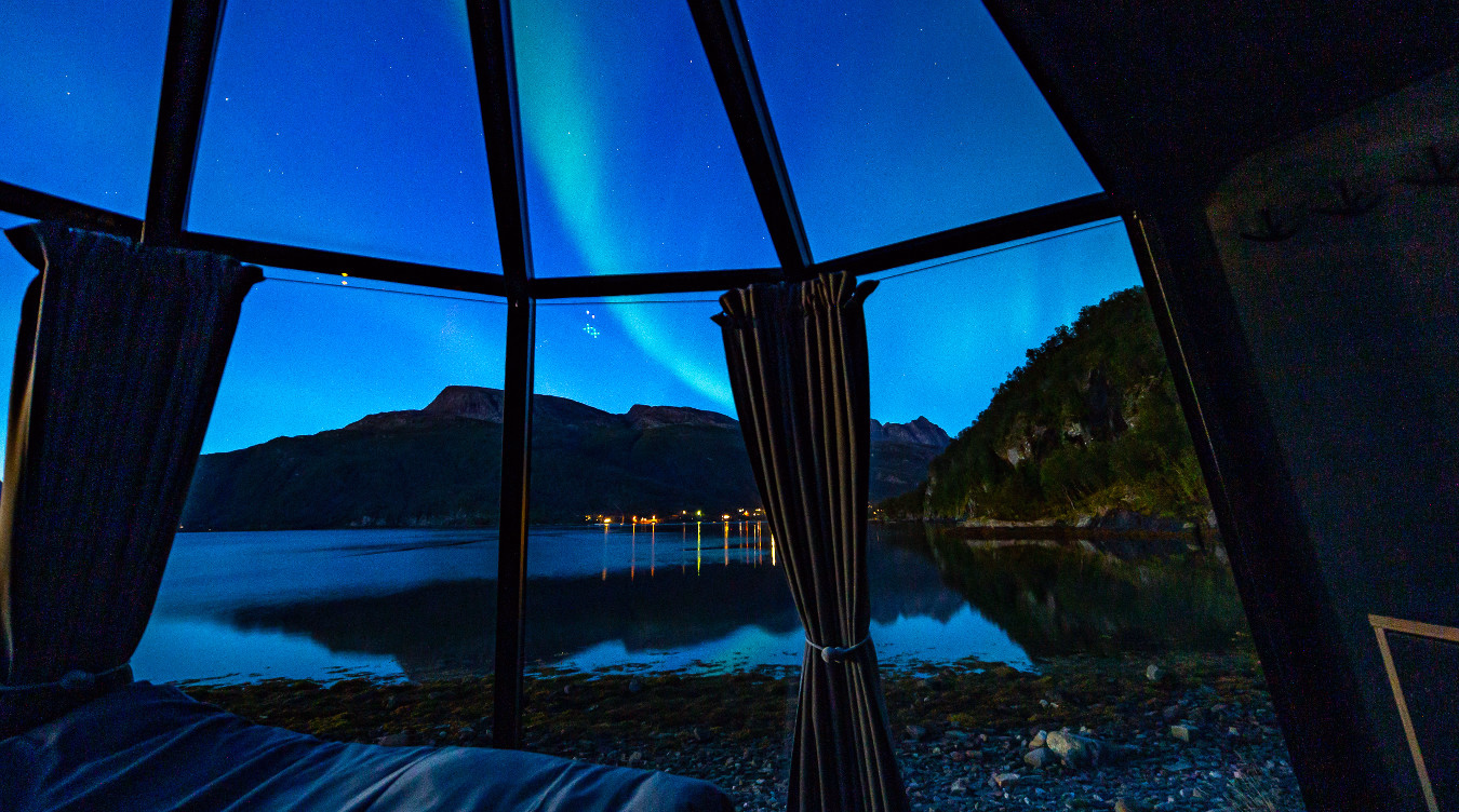 Nordlys over en innsjø og fjell sett gjennom et vindu fra et soverom