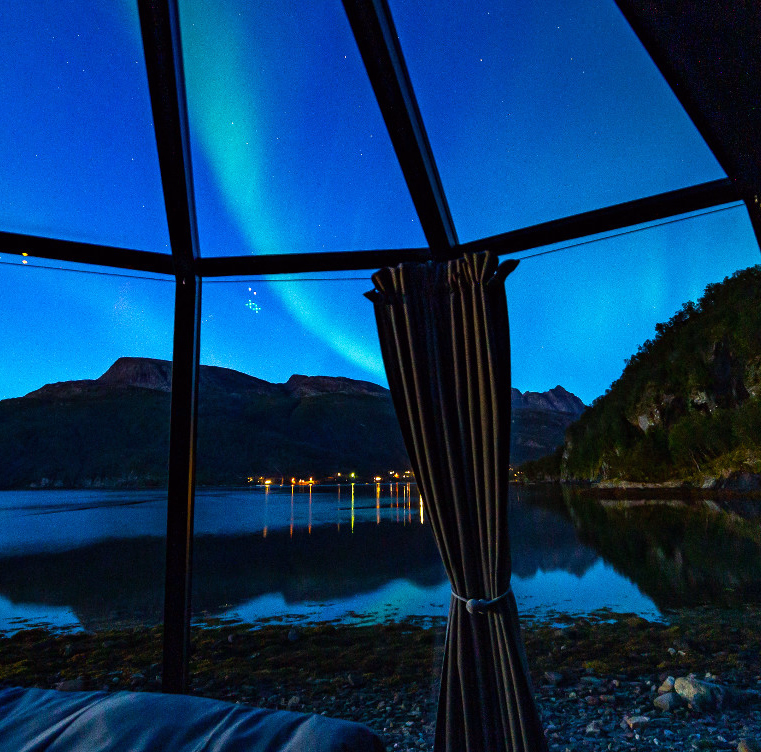 Nordlys over en innsjø og fjell sett gjennom et vindu fra et soverom