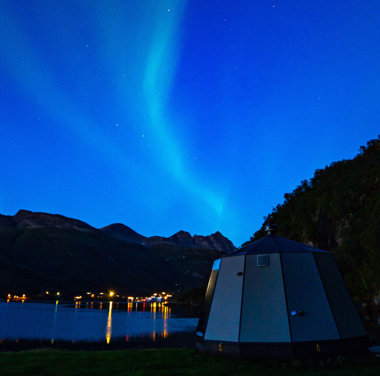 Grønt nordlys over en dome ved et vann med fjell i bakgrunnen