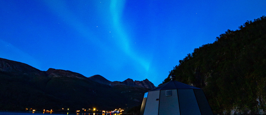 Grønt nordlys over en dome ved et vann med fjell i bakgrunnen