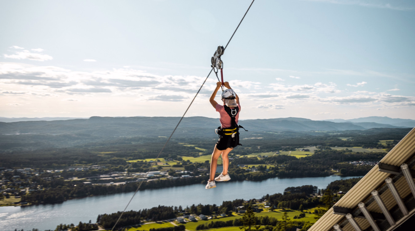 Person i rosa skjorte og hvit hjelm kjører zipline over en elv og et grønt landskap