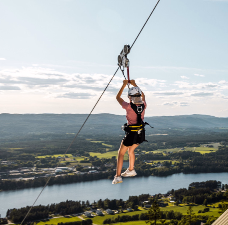 Person i rosa skjorte og hvit hjelm kjører zipline over en elv og et grønt landskap