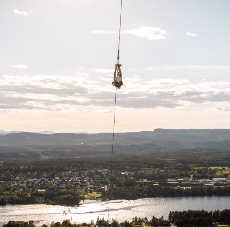 Person i strikkhopp henger opp ned over et landskap med elv og skog