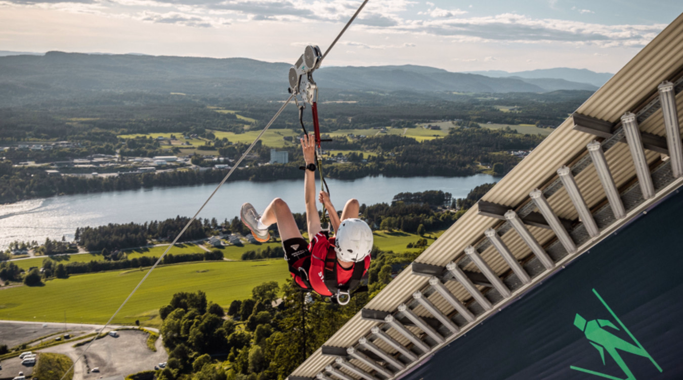 Person i rød skjorte og hvit hjelm kjører zipline over et landskap med en elv