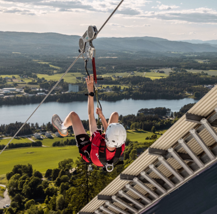 Person i rød skjorte og hvit hjelm kjører zipline over et landskap med en elv