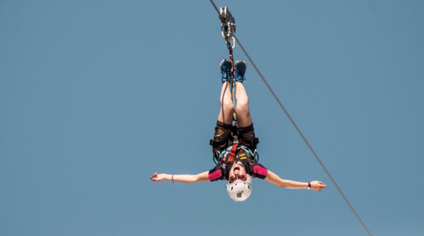 Person som henger opp ned i en zipline med utstrakte armer mot en blå himmel